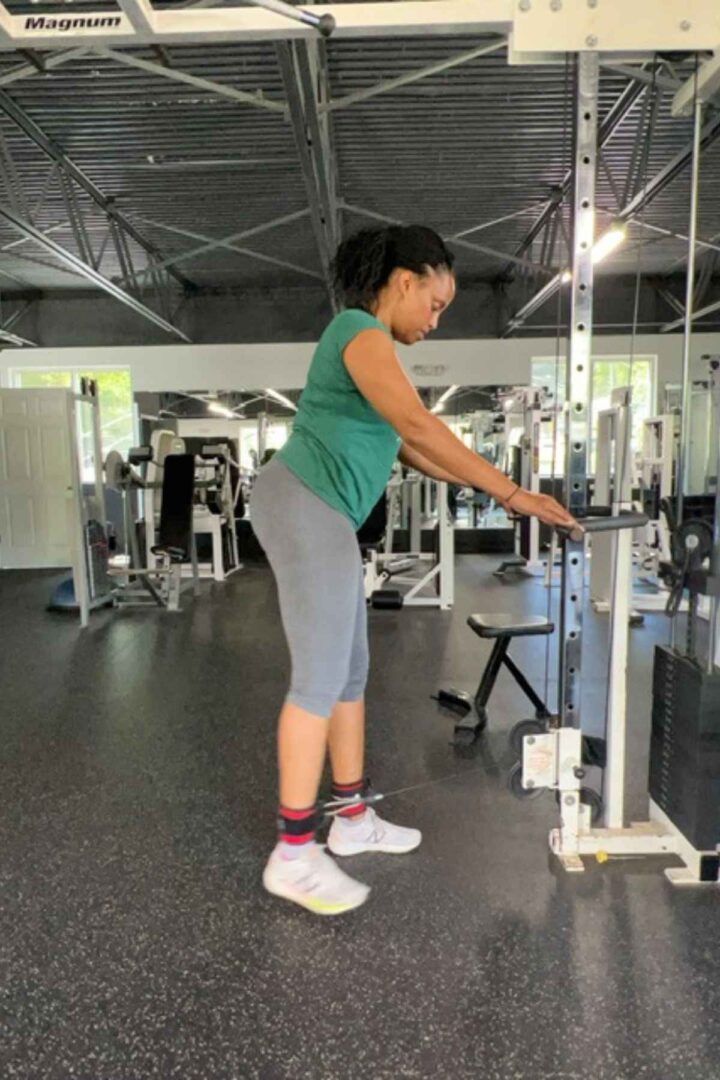 woman in green tshirt and gray leggings doing cable leg exercises with ankle strap