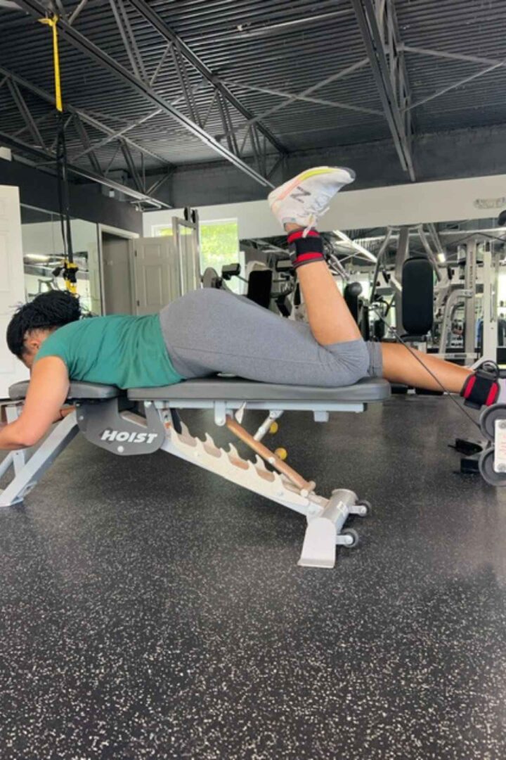 woman lying on a bench in a green tshirt and gray leggings lying hamstring cable leg exercise with ankle straps