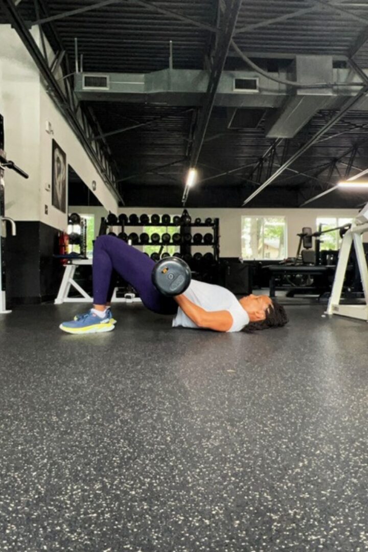 woman on the floor of gym in purple tights doing barbell glute bridges
